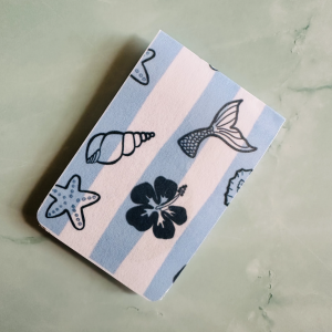 Marque-page magnétique "Ocean Vibes" : coquillage, queue de sirène, étoile de mer et hibiscus sur fond rayé bleu ciel et blanc, posé sur fond marbre vert