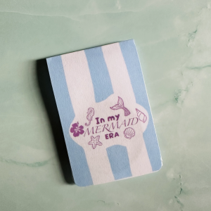 Marque-page magnétique "In My Mermaid Era" avec hippocampe, queue de sirène, coquillages et hibiscus en mauve sur fond rayé bleu ciel et rose poudré, posé sur fond marbre vert
