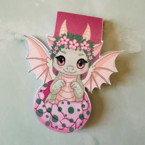 Marque-page magnétique die-cut "Lyra", bébé dragon vert kawaii couronné de fleurs roses émergeant d'un œuf rose tacheté, ailes déployées, posé sur fond marbre vert
