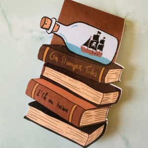 Marque-page magnétique die-cut représentant une pile de livres pirates "On Stranger Tides" et "L'île au Trésor" surmontée d'une bouteille à message avec navire pirate, posé sur fond marbre vert