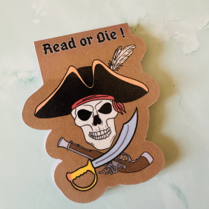 Marque-page magnétique die-cut "Read or Die!" : crâne de pirate avec chapeau à plume, bandeau rouge, sabre et pistolet croisés sur fond terracotta, posé sur fond marbre vert