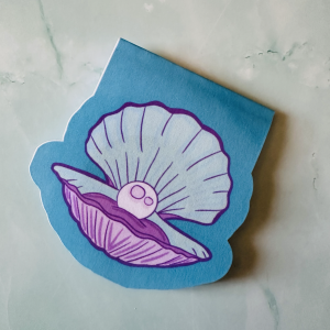 Marque-page magnétique die-cut "La Perle" : coquillage Saint-Jacques ouvert mauve et blanc avec une perle nacrée sur fond bleu ciel, posé sur fond marbre vert