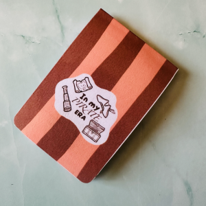 Marque-page magnétique "In My Pirate Era" avec carte au trésor, longue-vue, chapeau à plume et coffre en doodle brun sur fond rayé terracotta et saumon, posé sur fond marbre vert