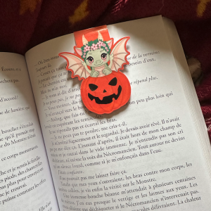 Marque-page magnétique die-cut "Happy Halloween – Lyra" : bébé dragonne verte kawaii couronnée de fleurs roses dans une citrouille Jack-o'-Lantern souriante, posé sur un livre ouvert