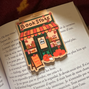 Marque-page magnétique die-cut "Book Store" : façade de librairie cosy automnale avec marquise rayée orange, renard endormi, hibou et feuilles d'érable, posé sur un livre ouvert