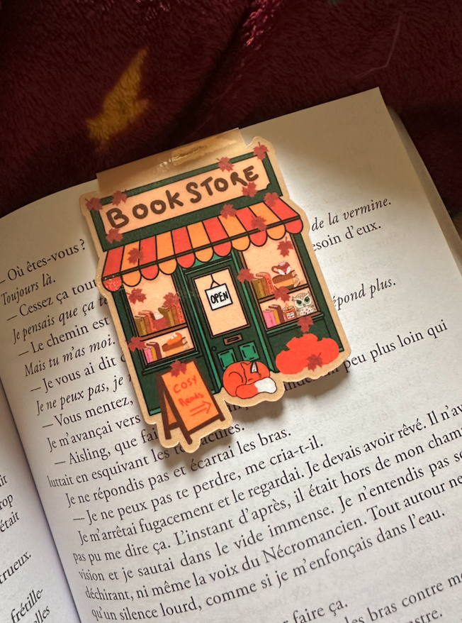 Marque-page magnétique die-cut "Book Store" : façade de librairie cosy automnale avec marquise rayée orange, renard endormi, hibou et feuilles d'érable, posé sur un livre ouvert