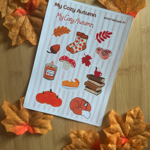 Planche de stickers "My Cozy Autumn" 12x17 cm : citrouilles, champignon, renard endormi, bougie Pumpkin Spice, cinnamon roll, pile de livres et feuilles d'automne sur fond rayé rose poudré, posée sur fond bois entouré de feuilles d'automne
