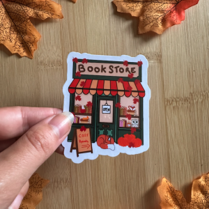 Sticker "Book Store" : façade de librairie cosy automnale avec marquise rayée orange, renard endormi, hibou et feuilles d'érable, tenu entre les doigts sur fond bois entouré de feuilles d'automne