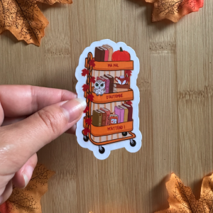 Sticker die-cut "Ma PAL d'Automne m'attend !" : chariot bibliothèque orange à trois étagères avec livres, citrouille, hibou kawaii et feuilles d'érable, tenu entre les doigts sur fond bois entouré de feuilles d'automne
