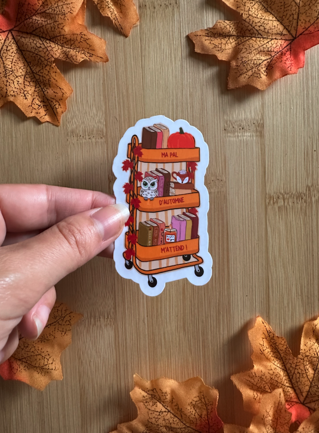 Sticker die-cut "Ma PAL d'Automne m'attend !" : chariot bibliothèque orange à trois étagères avec livres, citrouille, hibou kawaii et feuilles d'érable, tenu entre les doigts sur fond bois entouré de feuilles d'automne