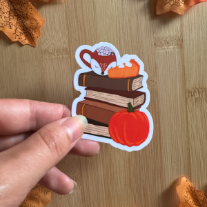 Sticker die-cut "Autumn Book Club" : pile de livres brun chocolat avec tasse renard marshmallows et citrouille orange, tenu entre les doigts sur fond bois entouré de feuilles d'automne