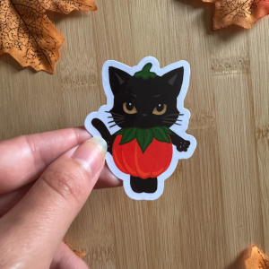 Sticker "Potichat d'Automne" : chat noir kawaii aux yeux dorés déguisé en citrouille orange, tenu entre les doigts sur fond bois entouré de feuilles d'automne