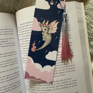Marque-page à pompon "Lyra" : recto dragonne verte kawaii couronnée de fleurs roses volant dans un ciel nocturne bleu avec nuages roses et étoiles, verso citation "Chaque page cache un souffle de magie" avec livre magique, pompon bicolore argent et rose dégradé, posé sur un livre ouvert