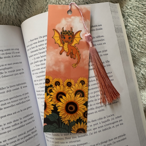 Marque-page à pompon "Maya" : recto dragonne orange kawaii couronnée de fleurs volant sur champ de tournesols fond pêche nuageux, verso citation "Books and Dragons Make the Best Summer" avec Maya et tournesols sur fond bordeaux, pompon rose dégradé, posé sur un livre ouvert